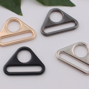 May include: Four triangle-shaped metal rings in gold, silver, black, and gunmetal. The rings have a small circle at the top and a long, thin rectangle in the middle.