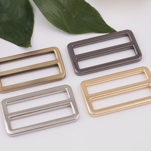May include: Four metal slider buckles in different finishes: gold, silver, antique brass, and black. Each buckle has a rectangular shape with two parallel slots.