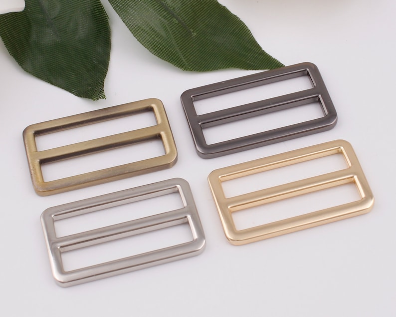 May include: Four metal slider buckles in different finishes: gold, silver, antique brass, and black. Each buckle has a rectangular shape with two parallel slots.