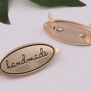 May include: Gold-toned metal oval tag with the word "handmade" engraved in black lettering. The tag has two prongs on the back for attaching to a product.