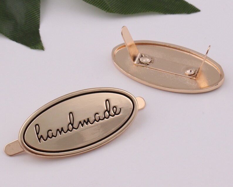 May include: Gold-toned metal oval tag with the word "handmade" engraved in black lettering. The tag has two prongs on the back for attaching to a product.