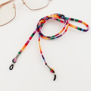 Puede incluir: Una cadena de gafas multirrayas colorida con extremos de metal plateado. La cadena est&aacute; hecha de tejido tejido y est&aacute; dise&ntilde;ada para sujetar las gafas de forma segura alrededor del cuello.
