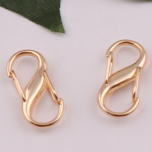 Pu&ograve; includere: Due chiusure metalliche a forma di S di colore dorato, anche conosciute come moschettoni, sono mostrate su uno sfondo bianco. Queste chiusure sono comunemente utilizzate per attaccare oggetti a borse, portachiavi o altri accessori.