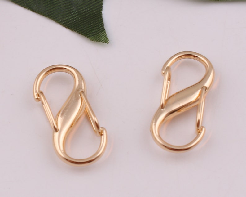 Pu&ograve; includere: Due chiusure metalliche a forma di S di colore dorato, anche conosciute come moschettoni, sono mostrate su uno sfondo bianco. Queste chiusure sono comunemente utilizzate per attaccare oggetti a borse, portachiavi o altri accessori.