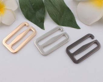 May include: Three metal bra strap hooks in gold, silver, and gunmetal colors. Each hook has a rectangular frame with a curved hook at one end and a central bar for strap adjustment. The hooks are displayed against a white background with floral accents.