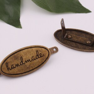 May include: Two oval-shaped metal tags with a brushed antique brass finish. One tag has the word "handmade" engraved on it. The other tag has two holes on the top and two holes on the bottom.