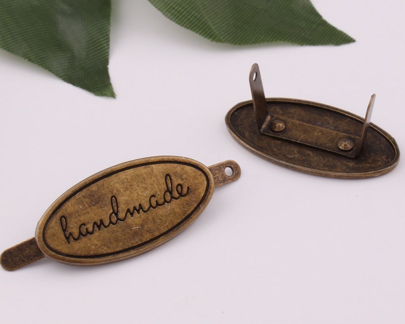 May include: Two oval-shaped metal tags with a brushed antique brass finish. One tag has the word "handmade" engraved on it. The other tag has two holes on the top and two holes on the bottom.