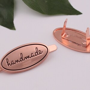 May include: A rose gold metal tag with the word "handmade" engraved in black lettering. The tag has a loop on one end and a clasp on the other end.