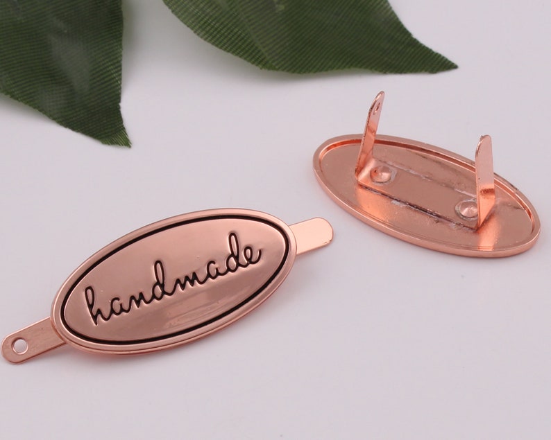 May include: A rose gold metal tag with the word "handmade" engraved in black lettering. The tag has a loop on one end and a clasp on the other end.
