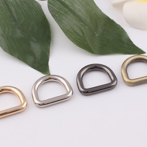May include: Four metal D-rings in different finishes: gold, silver, black, and antique brass. The rings are arranged in a row on a white surface.