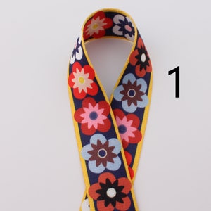Op de afbeelding: Een geel, blauw, rood en wit bloemenpatroon lint met een gele rand. Het lint is dubbelgevouwen en het nummer "1" is in wit op een witte achtergrond rechts van het lint geschreven.