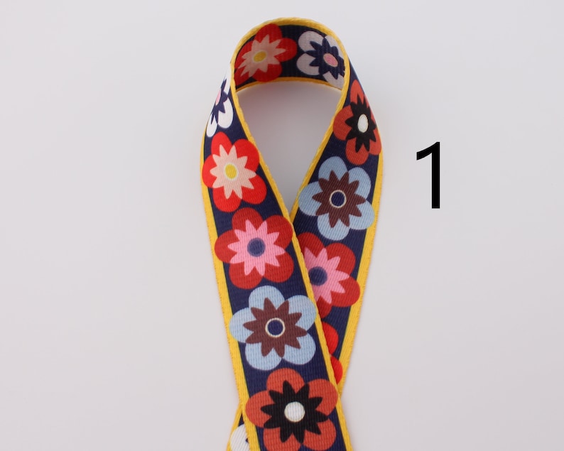 Op de afbeelding: Een geel, blauw, rood en wit bloemenpatroon lint met een gele rand. Het lint is dubbelgevouwen en het nummer "1" is in wit op een witte achtergrond rechts van het lint geschreven.