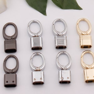 Könnte beinhalten: Acht Schlüsselringe aus Metall mit verschiedenen Oberflächen, darunter Gold, Silber und Schwarz. Jeder Ring hat eine quadratische Basis und eine Schlaufe oben.