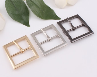 May include: Three metal belt buckles in gold, silver, and gunmetal gray. Each buckle has a square frame with a single bar for the belt strap. The buckles are displayed on a white surface with green leaves.