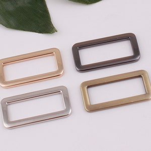 May include: Four metal rectangular rings in different finishes: gold, silver, brass, and black. The rings are all the same size and shape.