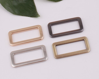 Puede incluir: Cuatro anillos rectangulares de metal en diferentes acabados: oro, plata, latón y negro. Los anillos son todos del mismo tamaño y forma.