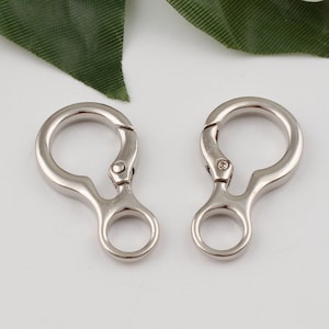 May include: Two silver metal swivel clasps with a small loop on each side. The clasps are open and ready to be attached to a bag or other item.