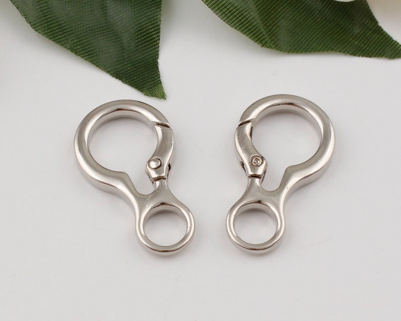 May include: Two silver metal swivel clasps with a small loop on each side. The clasps are open and ready to be attached to a bag or other item.