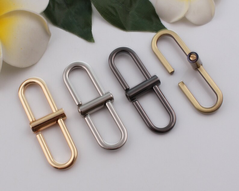 May include: Four metal D-ring clasps in gold, silver, black, and brass finishes. The clasps are oval shaped with a straight bar connecting the two loops.