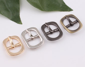 May include: Four metal belt buckles in different finishes: gold, silver, black, and antique brass. Each buckle has a rectangular shape with rounded corners and a center bar.