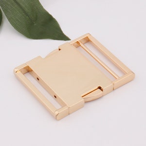May include: Gold metal buckle with a rectangular shape and two parallel bars. The buckle is designed to be used for belts, straps, or other similar applications.