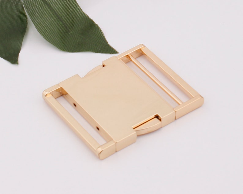 May include: Gold metal buckle with a rectangular shape and two parallel bars. The buckle is designed to be used for belts, straps, or other similar applications.