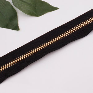May include: A black zipper with gold teeth. The zipper is lying flat on a white surface.