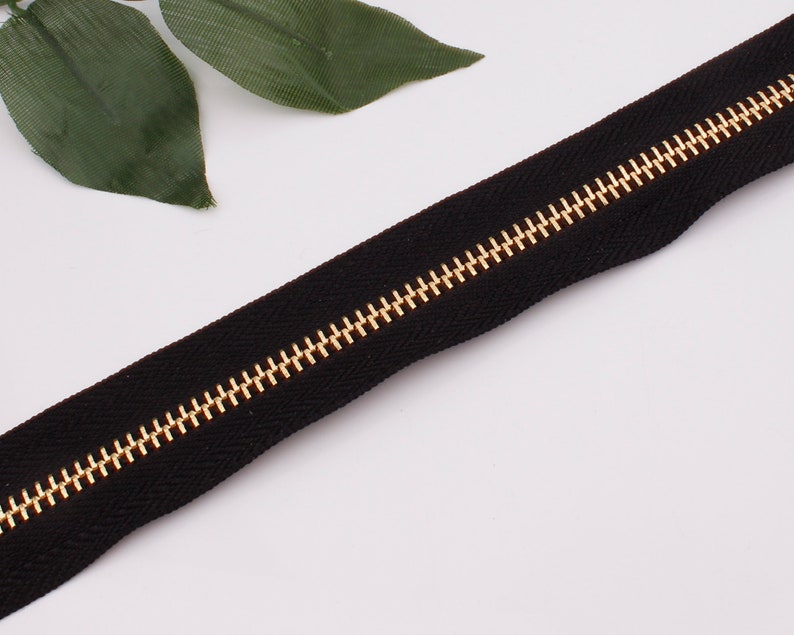 May include: A black zipper with gold teeth. The zipper is lying flat on a white surface.