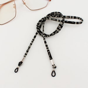Puede incluir: Cadena de gafas de patr&oacute;n blanco y negro con extremos de metal plateado. La cadena est&aacute; hecha de un material tejido y est&aacute; dise&ntilde;ada para sujetar las gafas de forma segura alrededor del cuello.