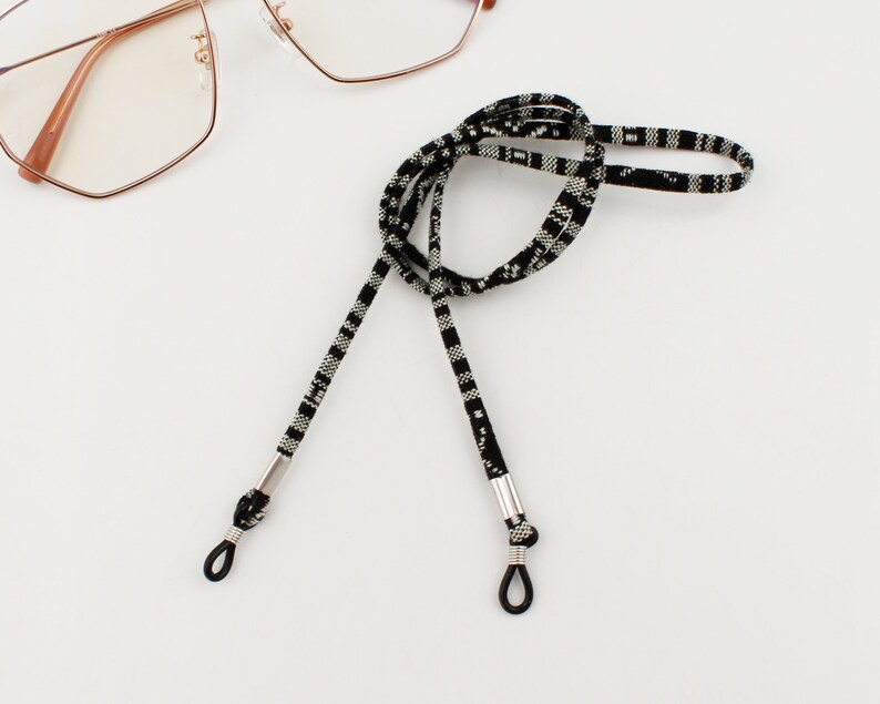 Puede incluir: Cadena de gafas de patr&oacute;n blanco y negro con extremos de metal plateado. La cadena est&aacute; hecha de un material tejido y est&aacute; dise&ntilde;ada para sujetar las gafas de forma segura alrededor del cuello.