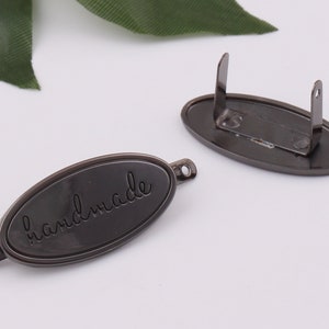 May include: A black metal tag with a rounded oval shape and a loop on the back. The tag has the word "handmade" engraved on the front.