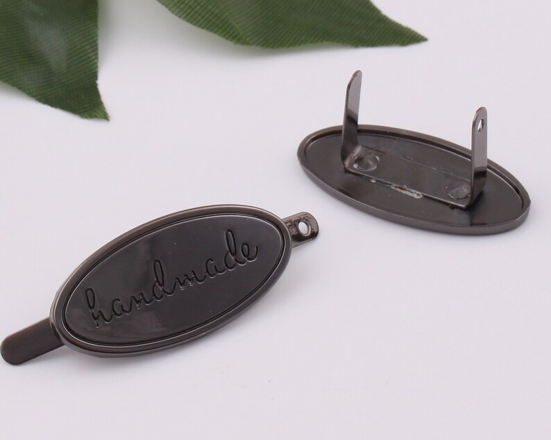 May include: A black metal tag with a rounded oval shape and a loop on the back. The tag has the word "handmade" engraved on the front.