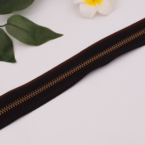 May include: A black zipper with a brown tape and gold metal teeth. The zipper is lying flat on a white surface.