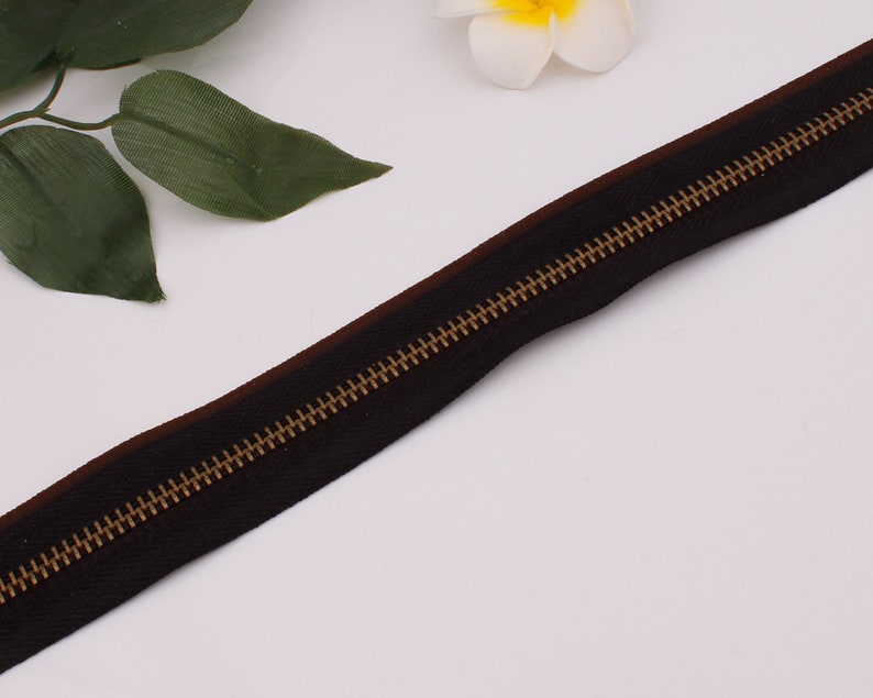 May include: A black zipper with a brown tape and gold metal teeth. The zipper is lying flat on a white surface.