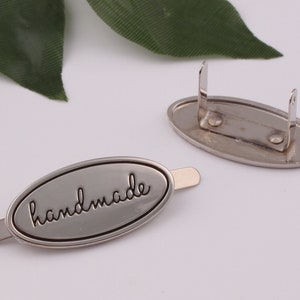 May include: Silver oval-shaped metal tag with the word "handmade" engraved in black lettering. The tag has a metal pin on the back for attaching to a product.