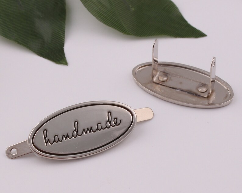 May include: Silver oval-shaped metal tag with the word "handmade" engraved in black lettering. The tag has a metal pin on the back for attaching to a product.