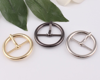 May include: Three metal belt buckles in gold, silver, and black. The buckles are round and have a simple design.