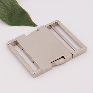 May include: Silver metal buckle with a rectangular shape and two slots for a strap. The buckle is designed to be used for belts, bags, or other accessories.