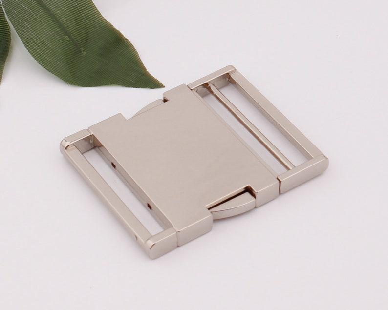 May include: Silver metal buckle with a rectangular shape and two slots for a strap. The buckle is designed to be used for belts, bags, or other accessories.