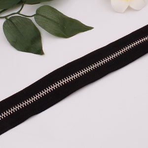 May include: A black zipper with a silver metal slider. The zipper is lying flat on a white surface.