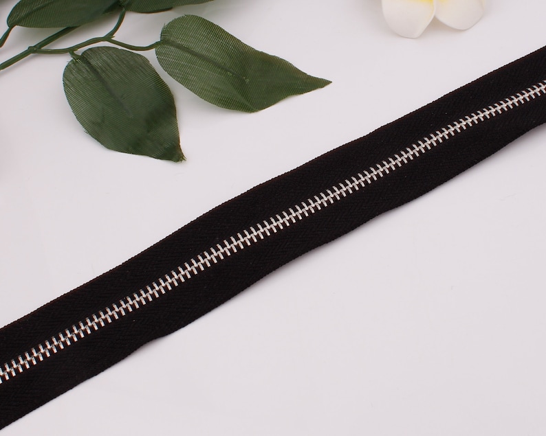 May include: A black zipper with a silver metal slider. The zipper is lying flat on a white surface.