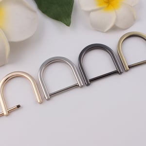 Op de afbeelding: Een verzameling metalen D-ringen in verschillende afwerkingen: goud, zilver, gunmetal en antiek messing. Deze halve cirkelvormige ringen hebben een rechte balk aan de onderkant. De ringen worden gebruikt voor het maken van tassen en andere knutselprojecten.