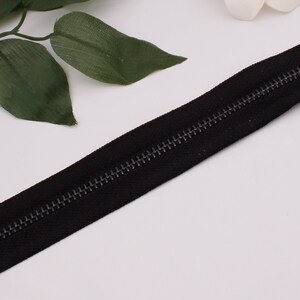 May include: A black zipper with a metal slider. The zipper is closed and the teeth are visible.