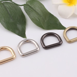 Puede incluir: Cuatro anillos en D de metal en diferentes acabados: dorado, plateado, negro y bronce. Los anillos están dispuestos en una fila sobre una superficie blanca.