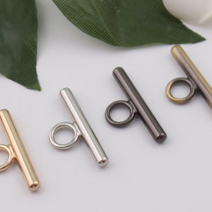 May include: Four metal toggle clasps in different finishes: gold, silver, black, and antique brass. Each clasp has a straight bar with a round loop at one end.