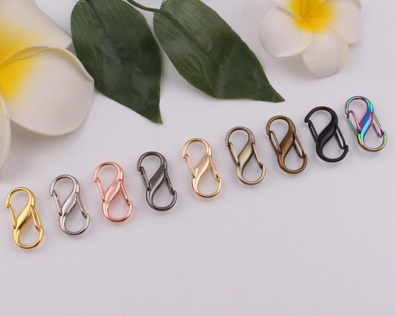Pu&ograve; includere: Assortimento di otto moschettoni in metallo in diverse finiture, tra cui oro, argento, oro rosa, nero, bronzo e arcobaleno.
