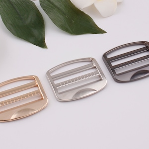 Puede incluir: Tres hebillas correderas metálicas en dorado, plateado y gris plomo. Cada hebilla tiene forma rectangular con bordes redondeados y una serie de barras y ranuras horizontales. Las hebillas se utilizan para ajustar correas en bolsos o accesorios.