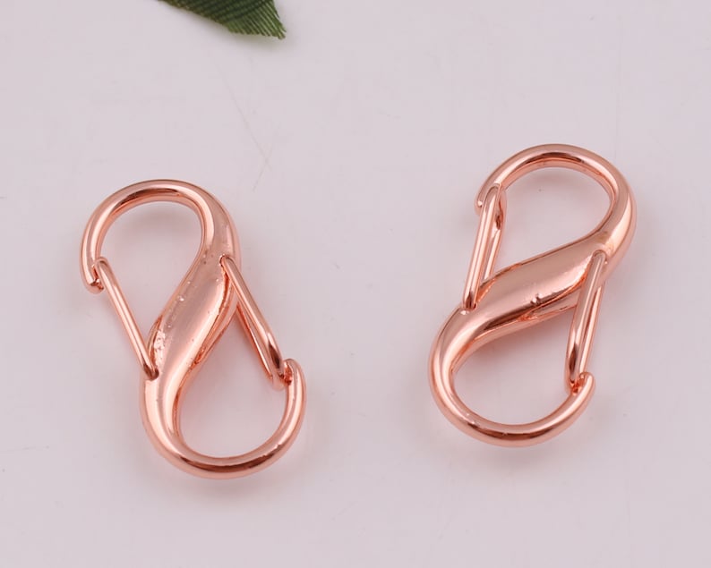 Pu&ograve; includere: Due chiusure a moschettone in metallo dorato rosa, ciascuna con un design a forma di otto.