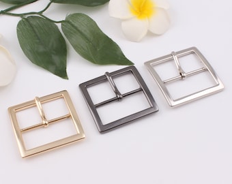 May include: Three metal belt buckles in gold, silver, and black. The buckles are square shaped with a single prong.