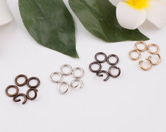 May include: An assortment of metal jump rings in various colors, including bronze, silver, gunmetal, and gold. Some rings are open, while others are closed. The jewelry findings are arranged on a white surface with green leaves and a white flower in the background.